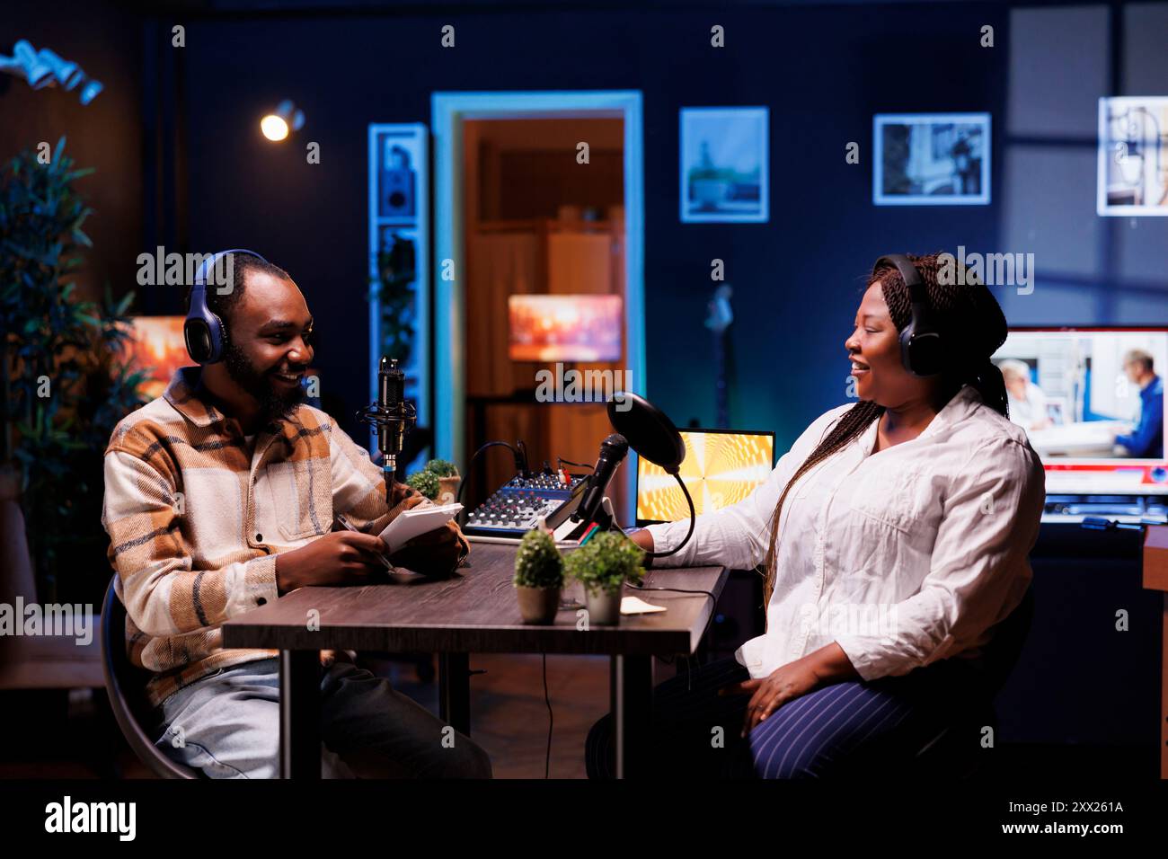 Afrikanische Podcaster, die im Heimstudio fröhlich diskutieren und verschiedene Themen mit Online-Zuhörern austauschen. Show Moderator mit Notizbuch interviewt weibliche Sozialitinnen und hat positive Interaktionen Stockfoto