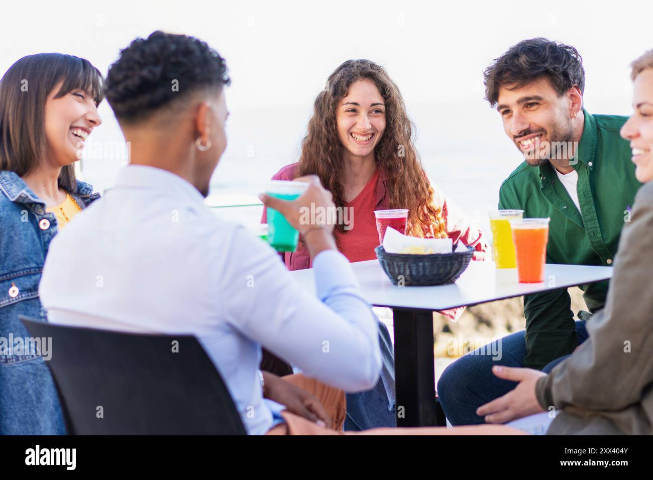 Fröhliche, abwechslungsreiche Gruppe junger Erwachsener, die Getränke und Snacks im Café im Freien teilt. Multikulturelle Freunde, die lachen und gesellig sind, vertreten die Incluusi Stockfoto