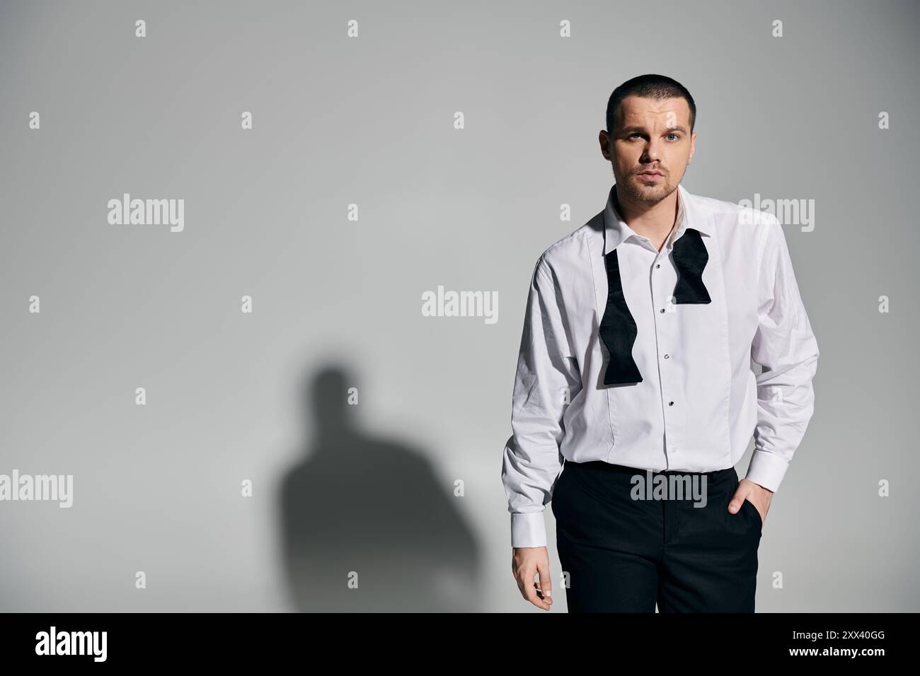 Ein Mann in weißem Hemd und schwarzem Bowtie steht selbstbewusst vor einem hellgrauen Hintergrund. Stockfoto