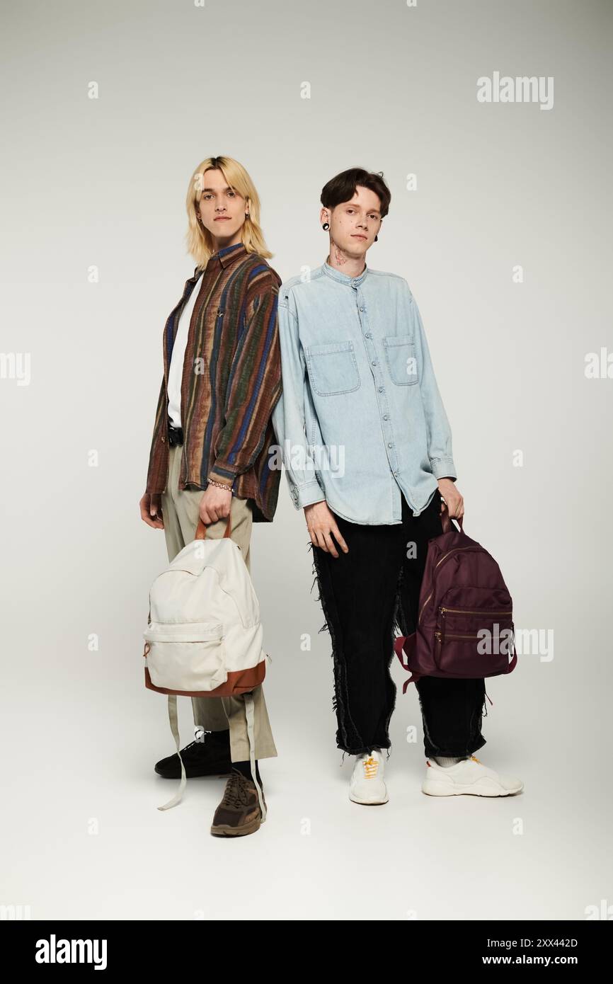 Zwei junge queere Studenten posieren zusammen in einem Studio, tragen legere Kleidung und halten Rucksäcke. Stockfoto