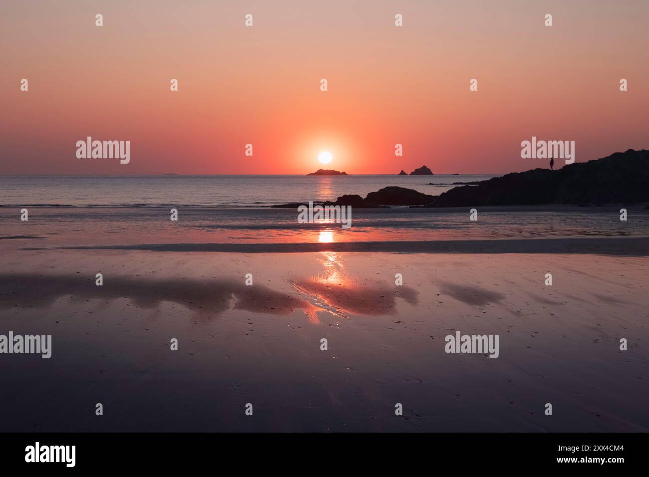 Leuchtender rosafarbener und oranger Sonnenuntergang über einem ruhigen Strand und Meer in Cornwall, Großbritannien, Sonnenuntergang am Horizont hinter Felsen Silhouetten im Ozean Stockfoto