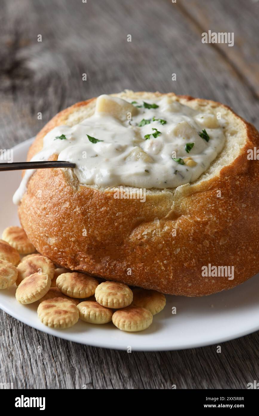 Vertikale Nahaufnahme von einem Brot Schüssel von New England Clam Chowder auf einem rustikalen Holztisch mit einem Löffel und Oyster Cracker. Stockfoto