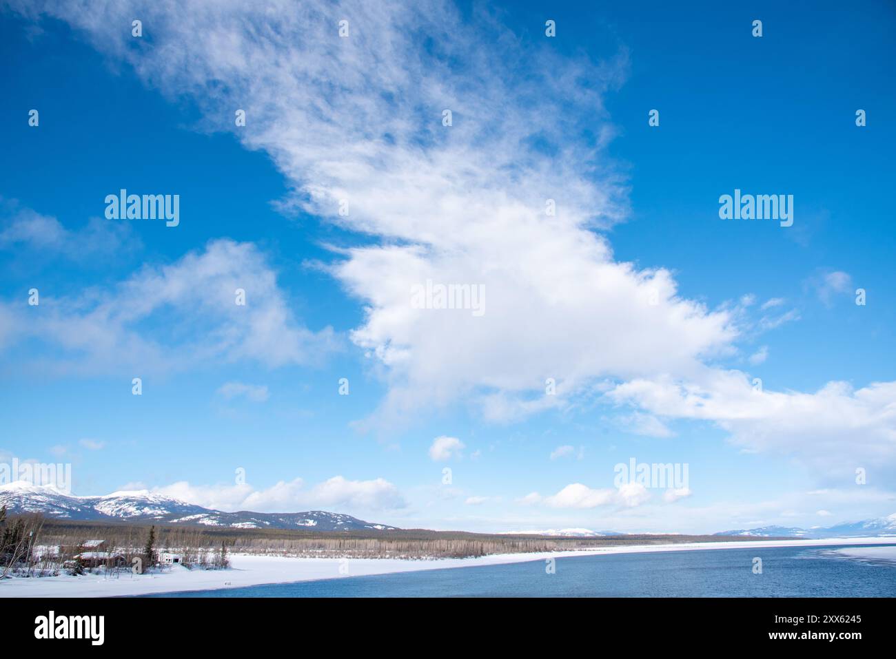 Ein blauer, heller Tag mit teilweise bewölkten Wolken und einem großen Fluss mit schneebedeckten Bergen und schneebedeckter Landschaft in Tagish, Yukon, Kanada Stockfoto