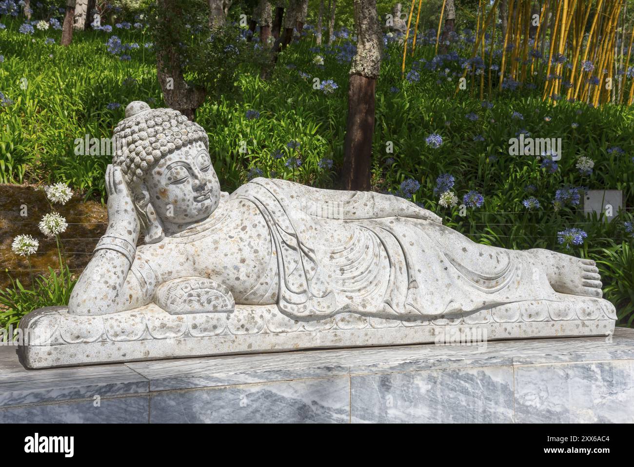 Stein-Buddha-Statue in liegender Position, umgeben von blühenden Pflanzen und Bäumen in einem sonnigen Park, Bacalhoa, Bacalhoa Buddha Eden, Quinta dos Lorid Stockfoto