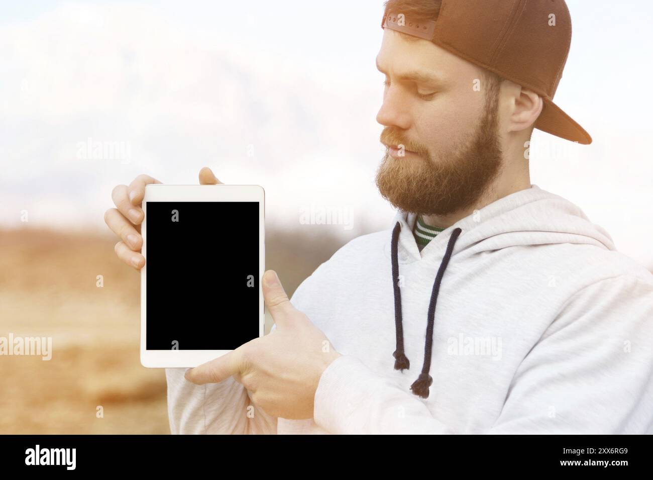 Nahaufnahme Porträt eines Hipsters in brauner Kappe im Freien hält einen weißen Tablet-pc in den Händen. Ein bärtiger Mann schaut sich die Tafel an. Hintergrundunschärfe Stockfoto