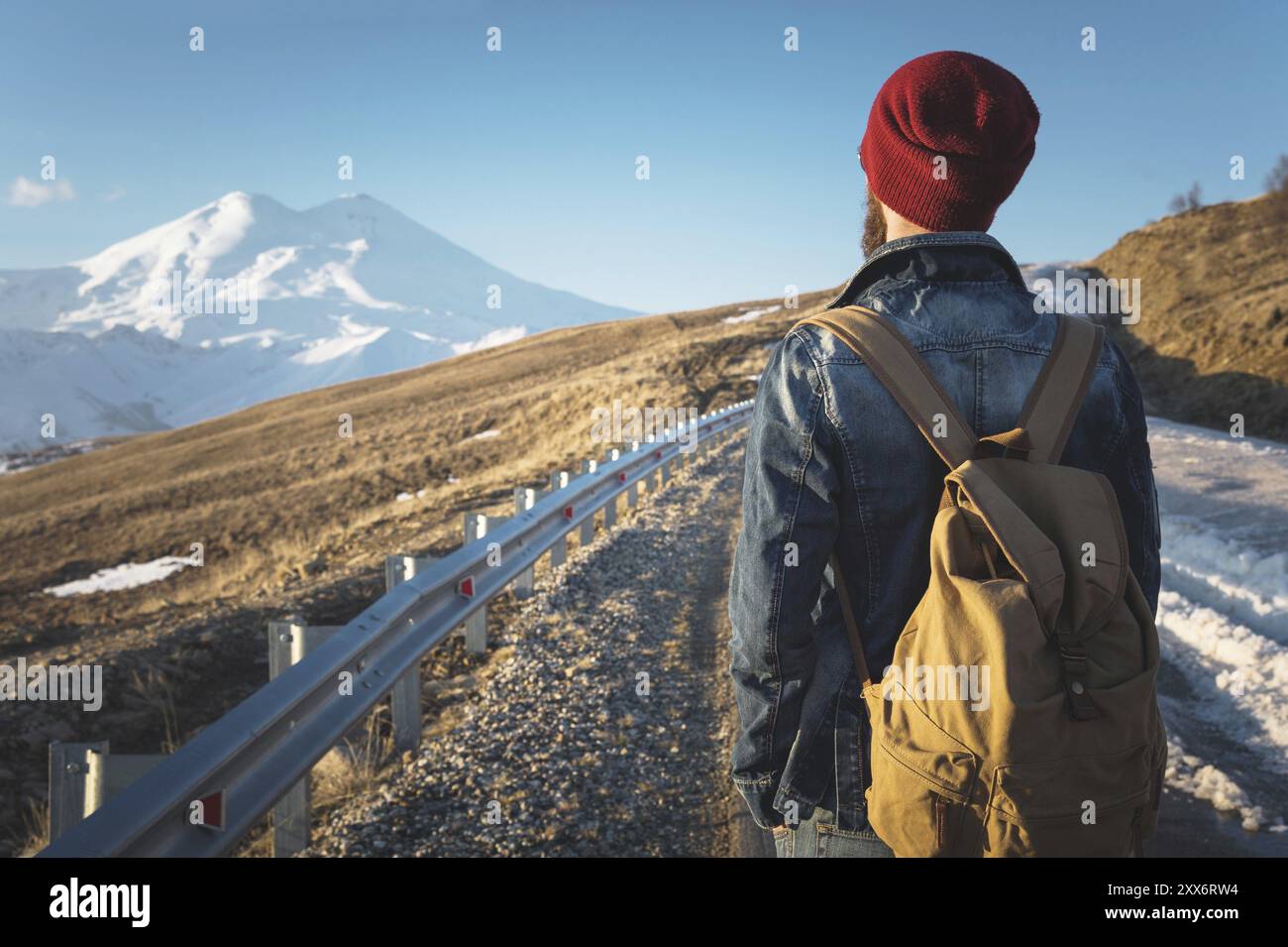 Ein bärtiger Hipster mit einem altmodischen Vintage-Rucksack mit Sonnenbrille und rotem Hut sowie Jeansjacke und Jeans steht auf einem Landasphalt Stockfoto