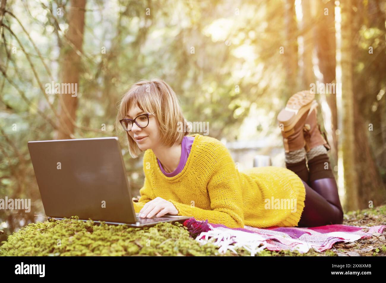 Getöntes Bild eines freiberuflichen Mädchenporträts in einem gelben Pullover und einer Brille, der nachdenklich in der Natur in einem Nadelwald auf den Laptop-Bildschirm blickt Stockfoto