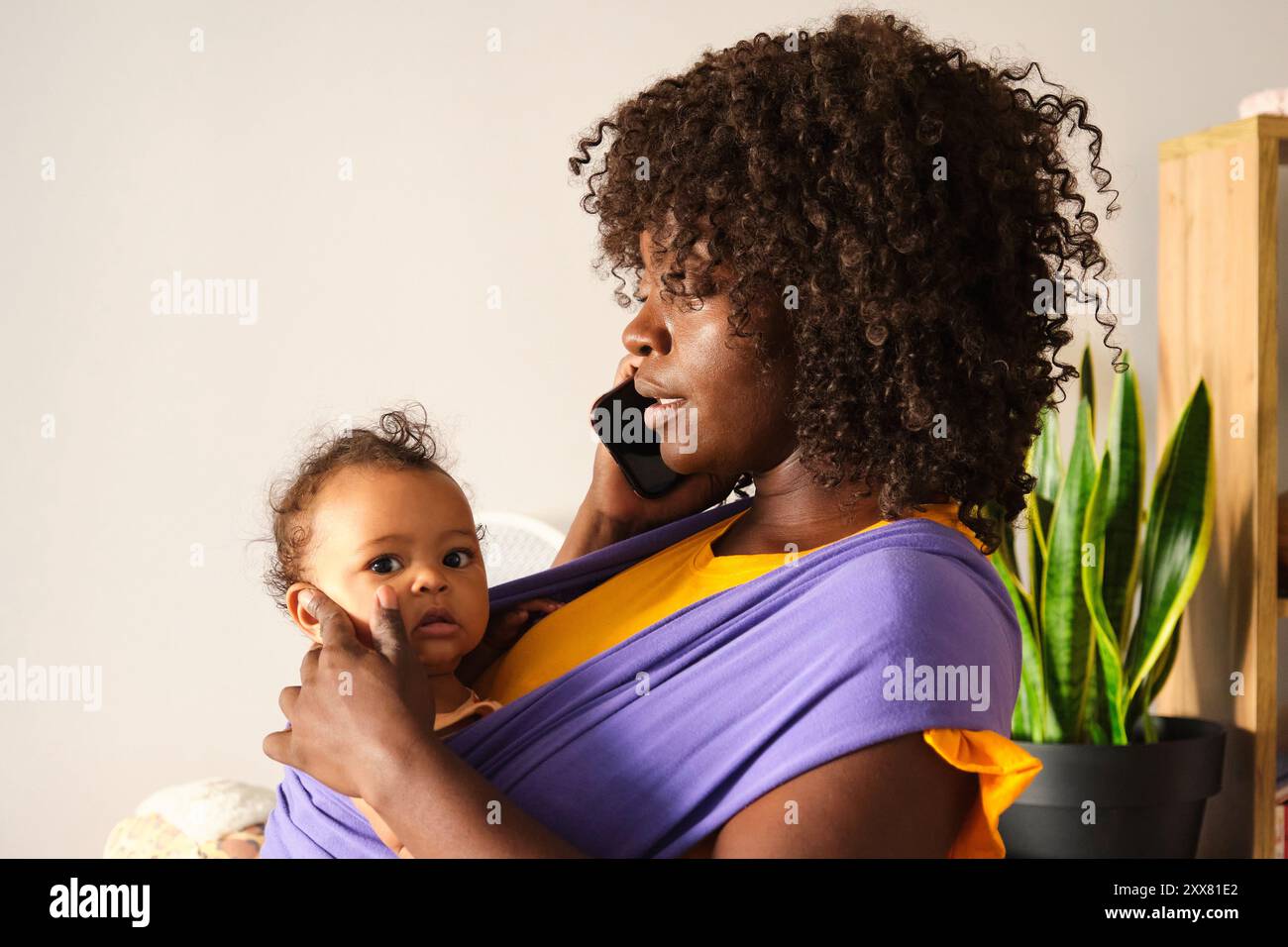 Mutter, die von zu Hause aus arbeitet, hält das Baby mit einem Hebegurt fest, der am Telefon spricht Stockfoto