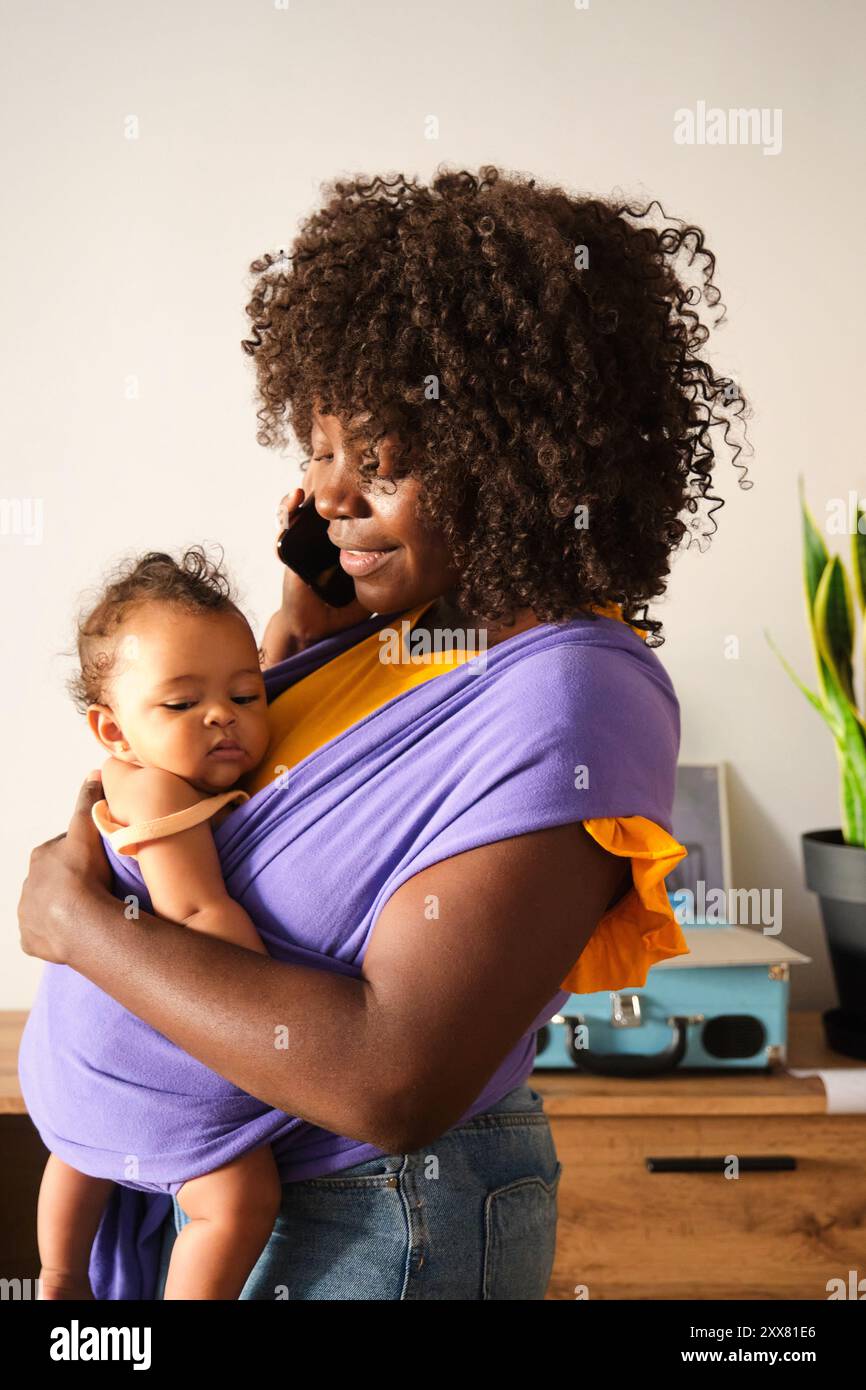Mutter, die von zu Hause aus arbeitet, hält das Baby mit einem Hebegurt fest, der am Telefon spricht Stockfoto