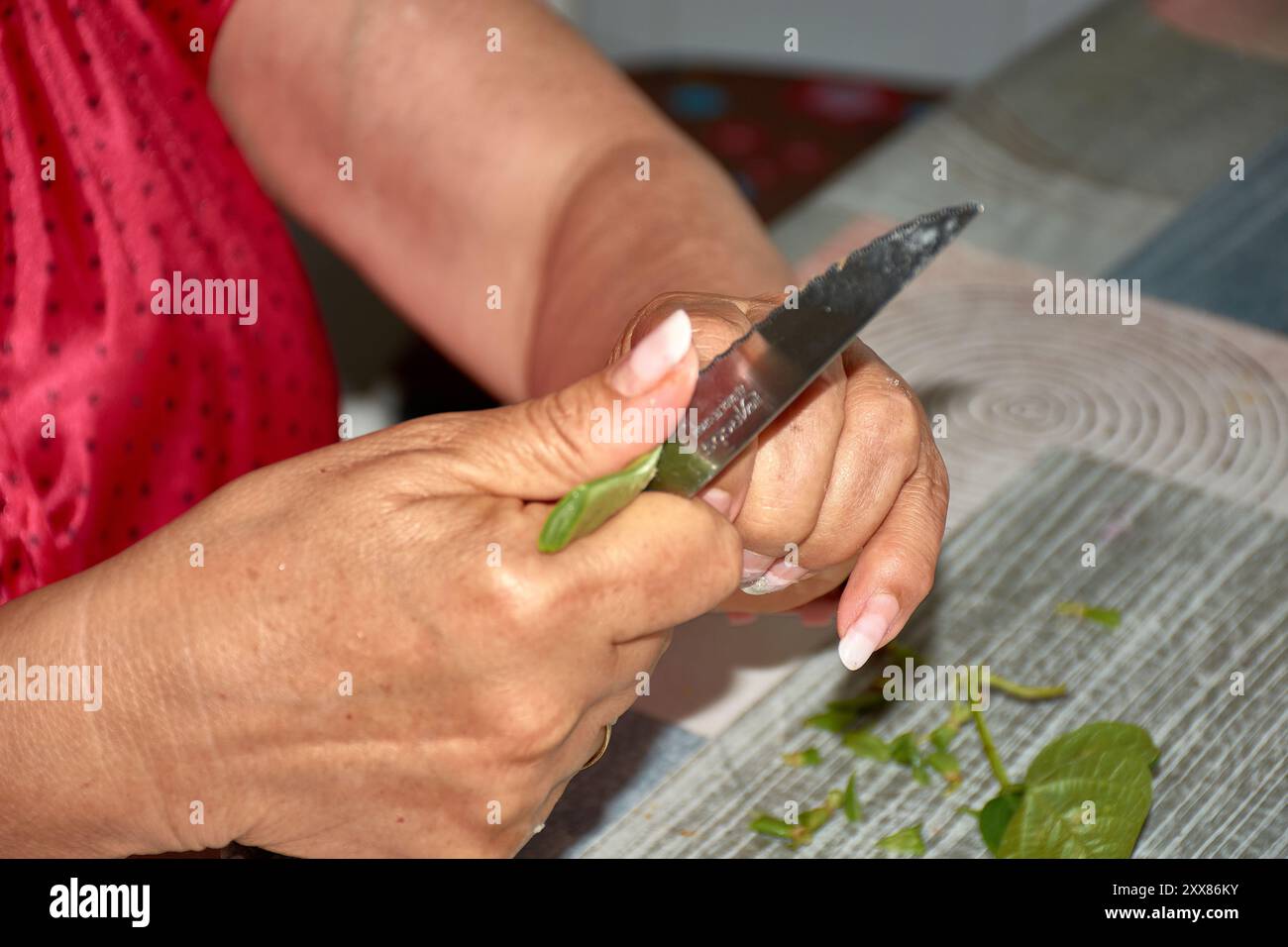 Die Hände einer älteren Frau, während sie vorsichtig grüne Bohnen mit einem Messer schneidet, um sie zum Kochen vorzubereiten. Die verwitterten, aber geschickten Hände, gekennzeichnet mit b Stockfoto