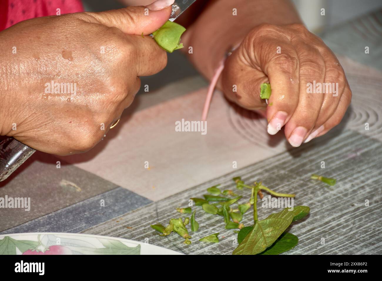 Die Hände einer älteren Frau, während sie vorsichtig grüne Bohnen mit einem Messer schneidet, um sie zum Kochen vorzubereiten. Die verwitterten, aber geschickten Hände, gekennzeichnet mit b Stockfoto