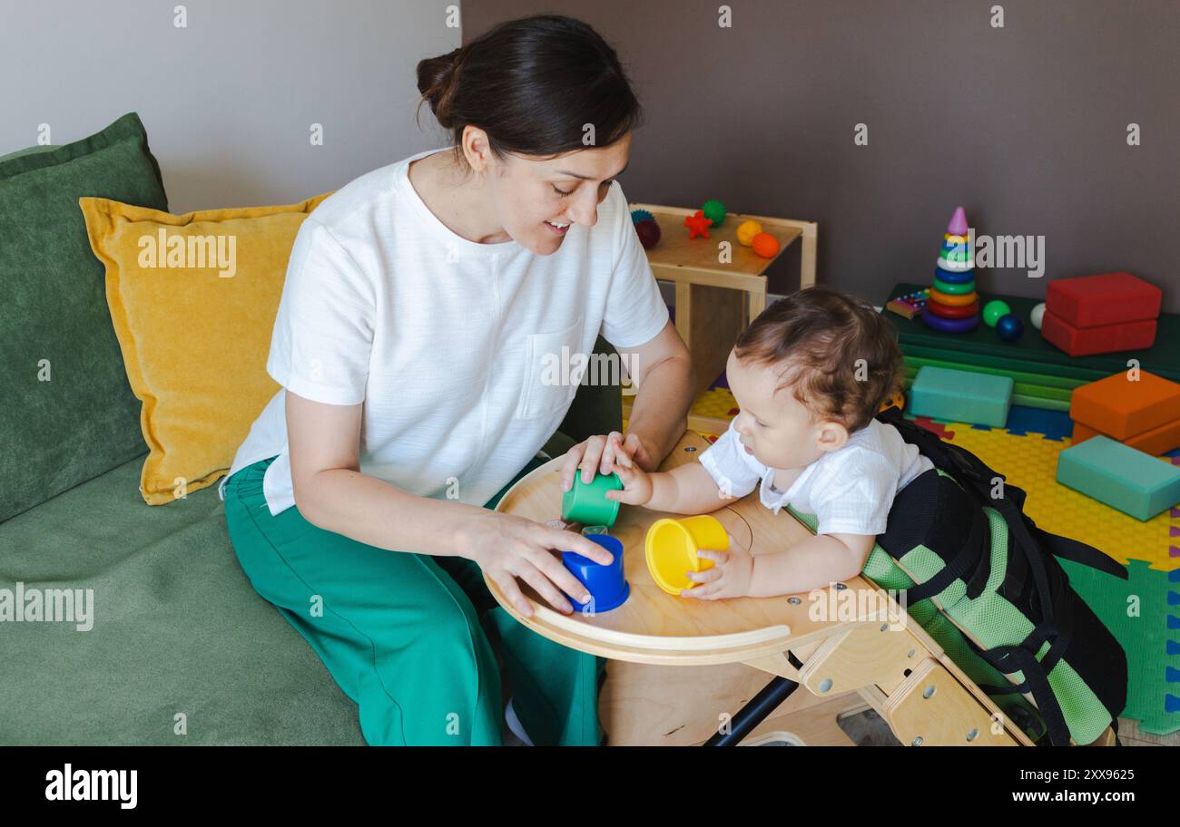 Männliches Kind mit Erkrankungen des Bewegungsapparates nimmt eine Rehabilitation mit einem Physiotherapeuten Teil, wobei farbenfrohe Becher zur Entwicklung der Fähigkeiten verwendet werden Stockfoto