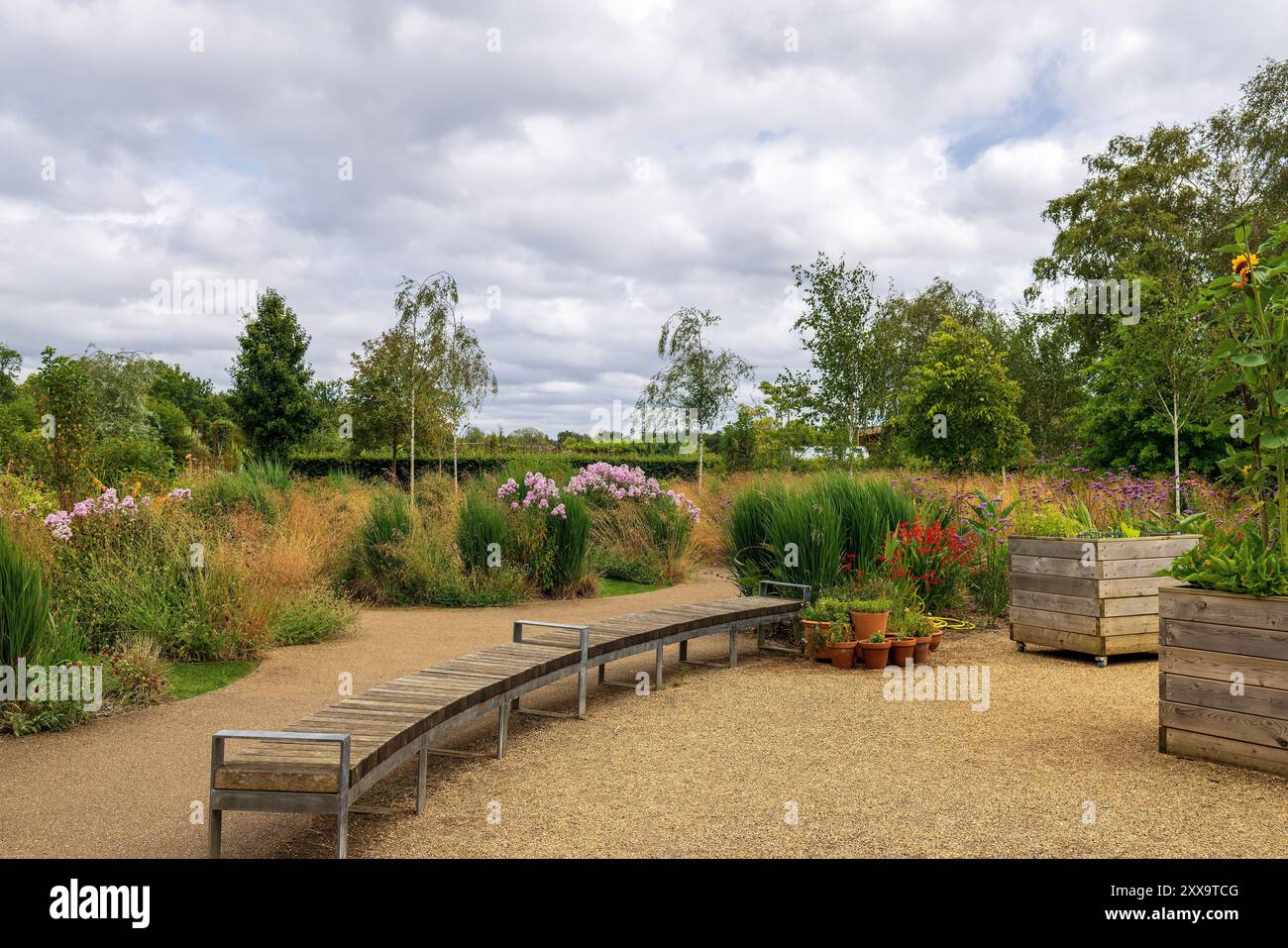 Lange gekrümmte Holzbank in einem Park mit erhöhten Blumenbeeten und Kiesweg. Stockfoto