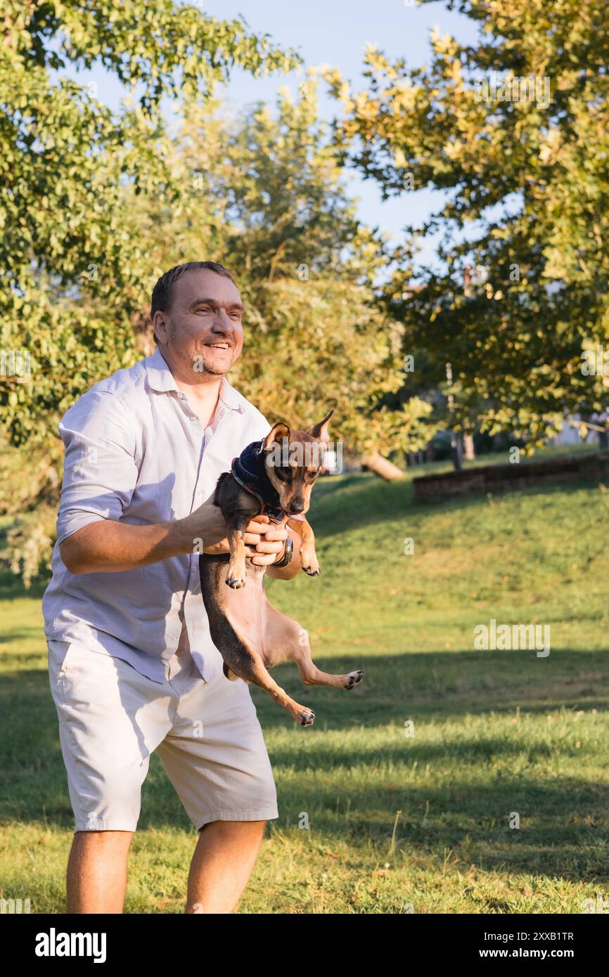 Ein lächelnder Mann mittleren Alters spaziert und spielt mit einem kleinen Hund im öffentlichen Park Stockfoto