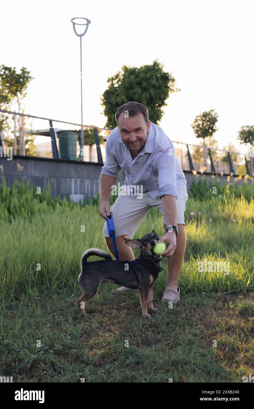 Ein lächelnder Mann mittleren Alters spaziert und spielt mit einem kleinen Hund im öffentlichen Park Stockfoto