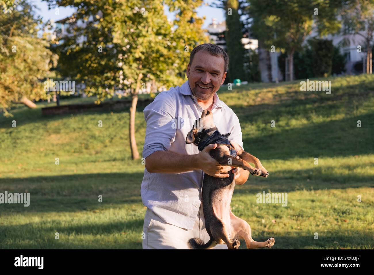 Ein lächelnder Mann mittleren Alters spaziert und spielt mit einem kleinen Hund im öffentlichen Park Stockfoto