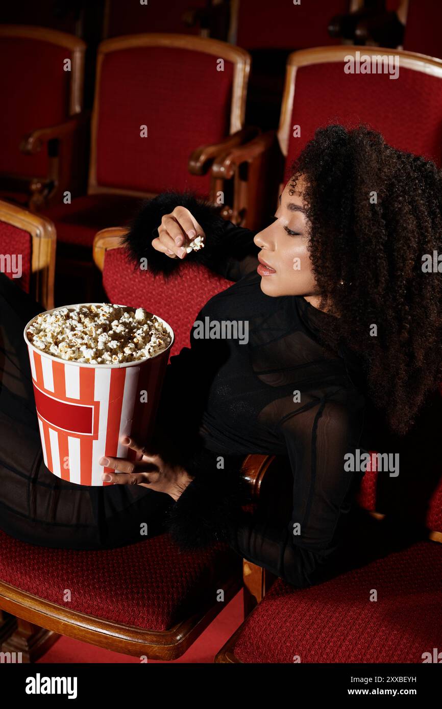 Eine stilvolle Frau in einem Kino, vertieft in den Film, genießt einen Eimer Popcorn. Stockfoto