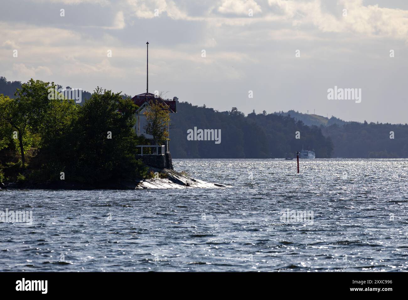 Ein ruhiger Blick auf den See mit einem kleinen Haus an einem felsigen Ufer, umgeben von Bäumen. Das Wasser reflektiert den bewölkten Himmel, und ein weit entferntes Boot ist zu sehen Stockfoto