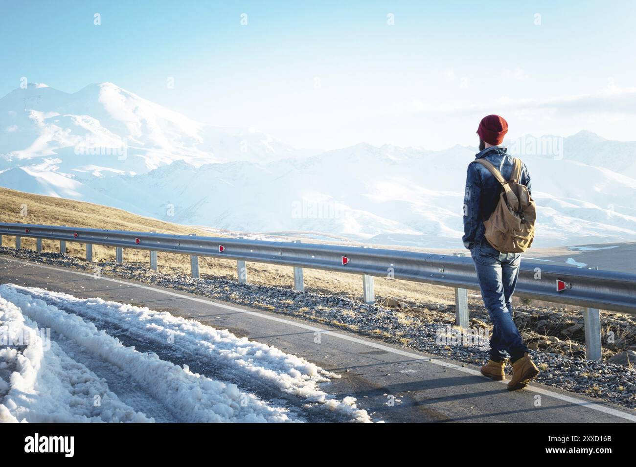 Ein stylischer bärtiger Hipster in Sonnenbrille mit einem Vintage-Rucksack spaziert an einem sonnigen Tag entlang der Asphaltstraße. Das Konzept des Anhalter- und Wanderns Stockfoto