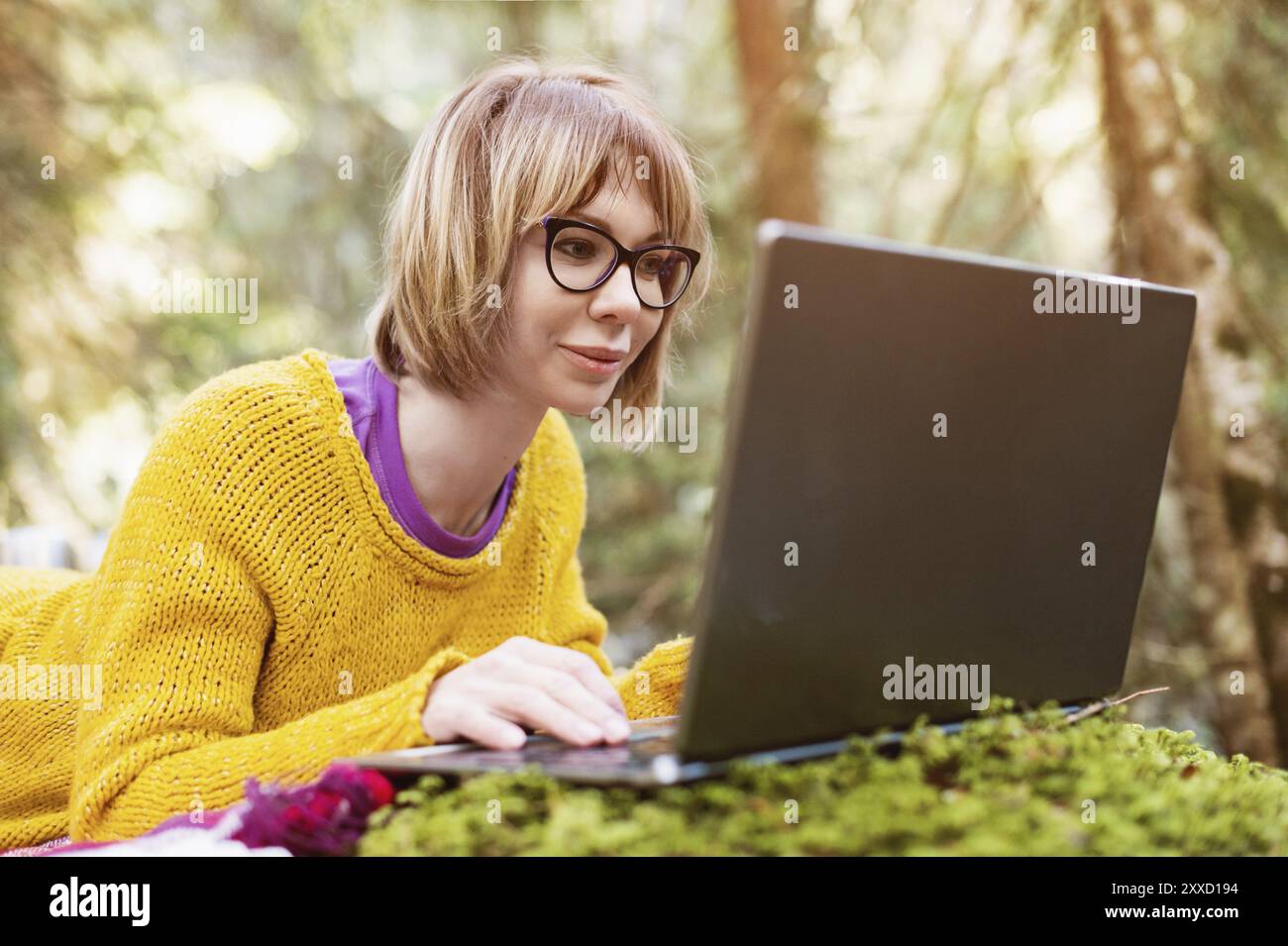 Getöntes Bild eines freiberuflichen Mädchenporträts in einem gelben Pullover und einer Brille, der nachdenklich in der Natur in einem Nadelwald auf den Laptop-Bildschirm blickt Stockfoto