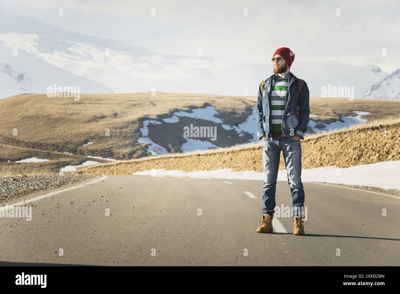 Ein stylischer bärtiger Hipster in Sonnenbrille mit einem Vintage-Rucksack steht an einem sonnigen Tag auf einem Landstraßenasphalt. Das Konzept des Anhalter- und Wanderns Stockfoto