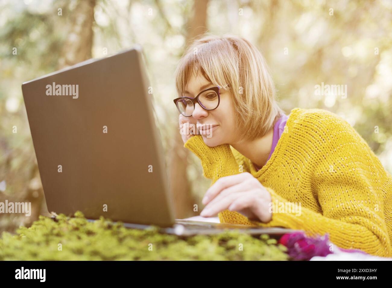 Getöntes Bild eines freiberuflichen Mädchenporträts in einem gelben Pullover und einer Brille, der nachdenklich in der Natur in einem Nadelwald auf den Laptop-Bildschirm blickt Stockfoto