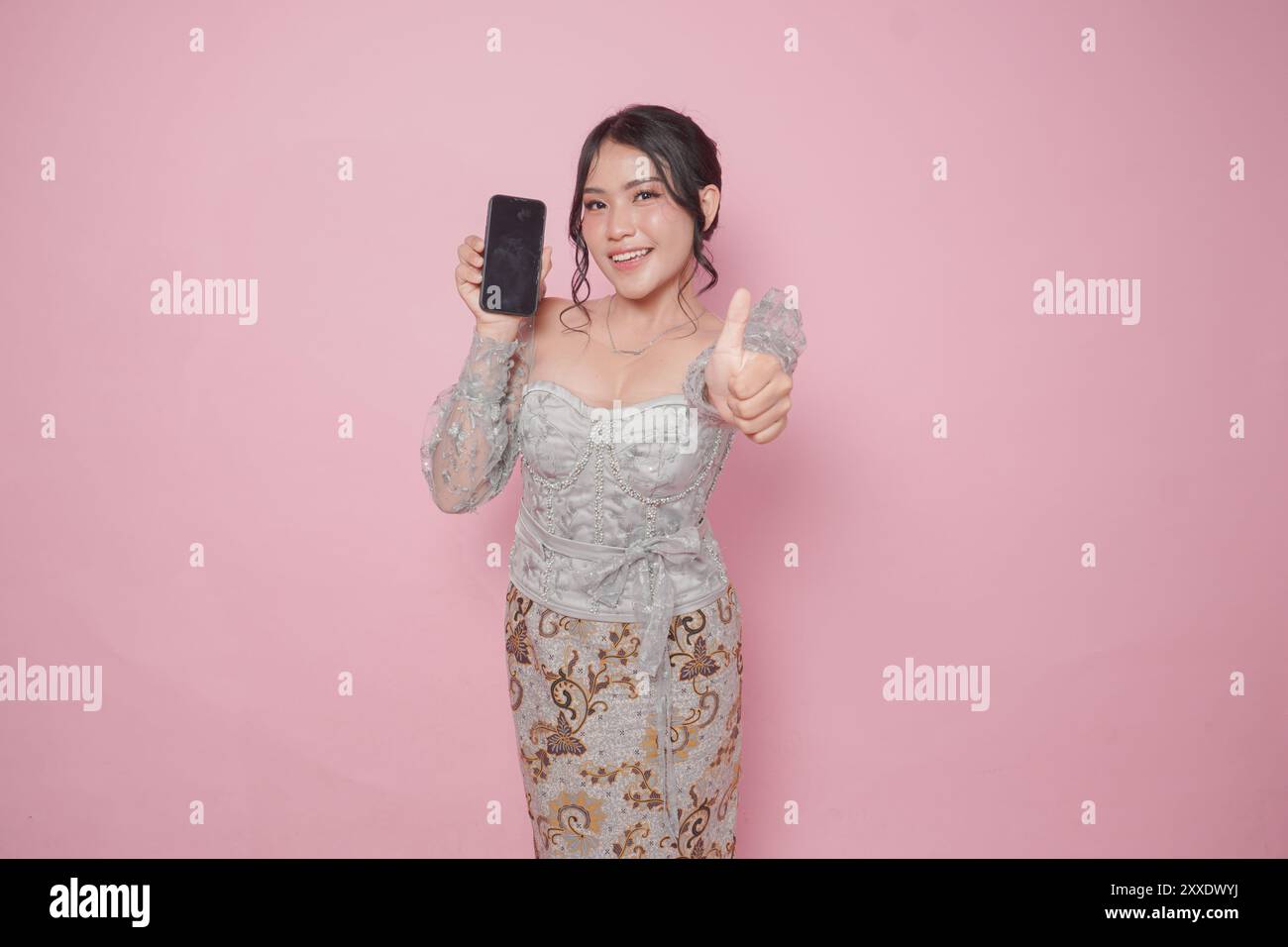 Die aufgeregte indonesische Frau in modernem Kebaya-Kleid gibt die Daumen hoch und zeigt auf ihrem Handy einen Kopierraum, isoliert durch einen rosa Hintern Stockfoto