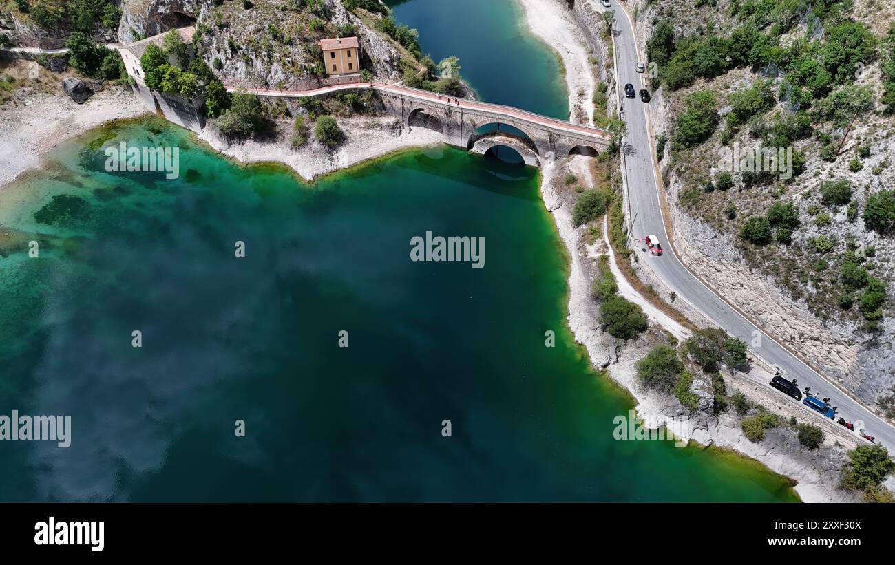 Das ruhige Wasser eines Sees glitzert unter der Sonne, mit einer rustikalen Brücke, die felsige Ufer verbindet, und einem malerischen Gebäude inmitten von Grün. Stockfoto
