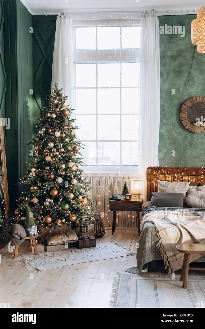 Das Schlafzimmer ist weihnachtlich dekoriert mit einem Weihnachtsbaum mit grünen, goldenen, kupfernen und silbernen Kugeln. Großes Fenster, hölzernes Kopfteil Bett mit Feenlichtern Stockfoto