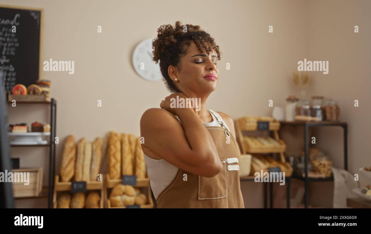 Junge hispanische Frau in einer Bäckerei, die ihren Hals vor Schmerzen hält, umgeben von frischem Brot in einem Innenladen Stockfoto