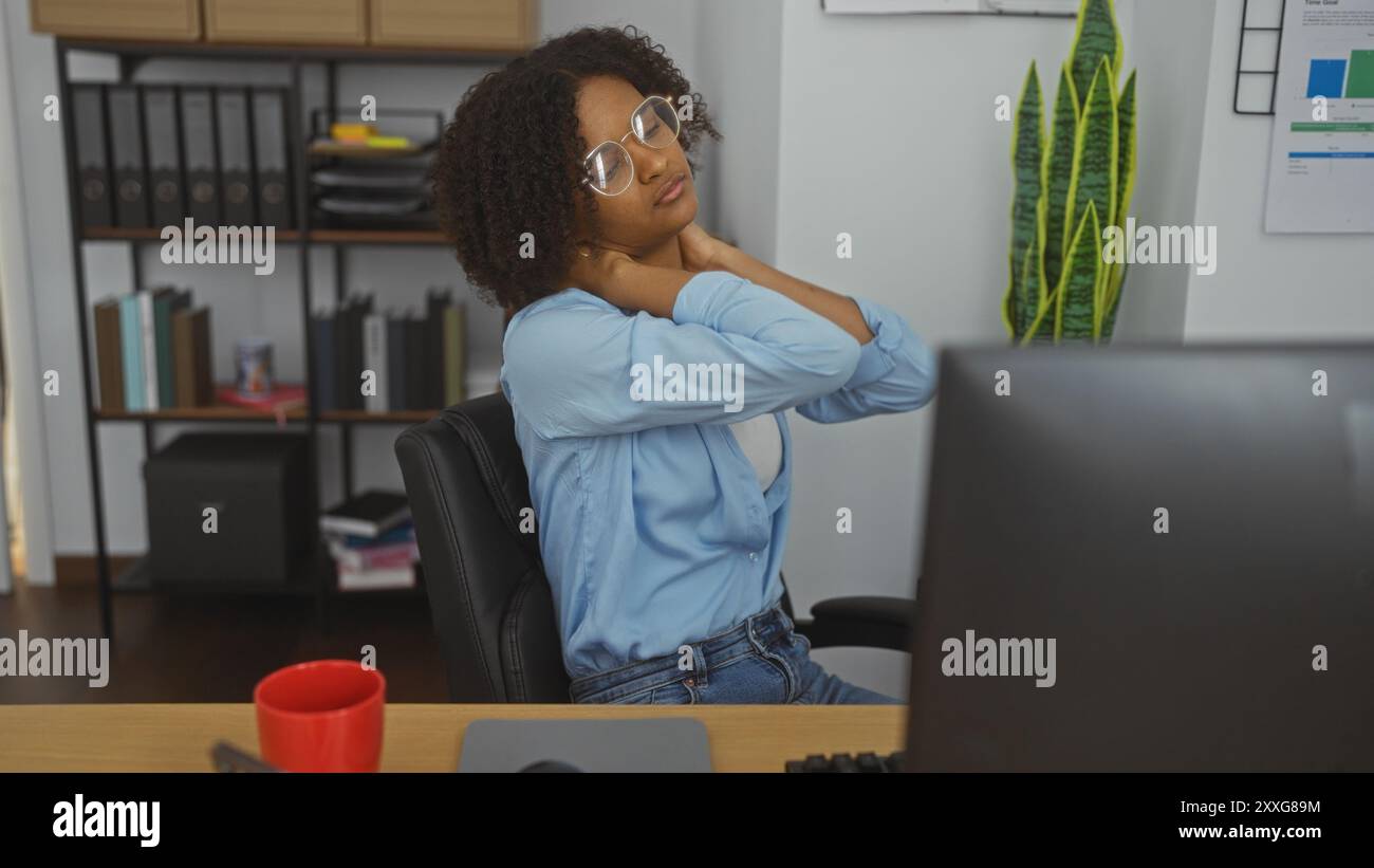 Lockige junge Frau mit Brille, die Nackenschmerzen in einer Büroumgebung mit Computer und roter Tasse auf dem Schreibtisch spürt Stockfoto