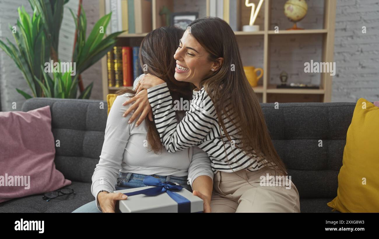 Eine lächelnde Frau umarmt ihre Tochter, die ein Geschenk in einem gemütlichen Wohnzimmer hält, das Liebe und familiäre Wärme symbolisiert. Stockfoto
