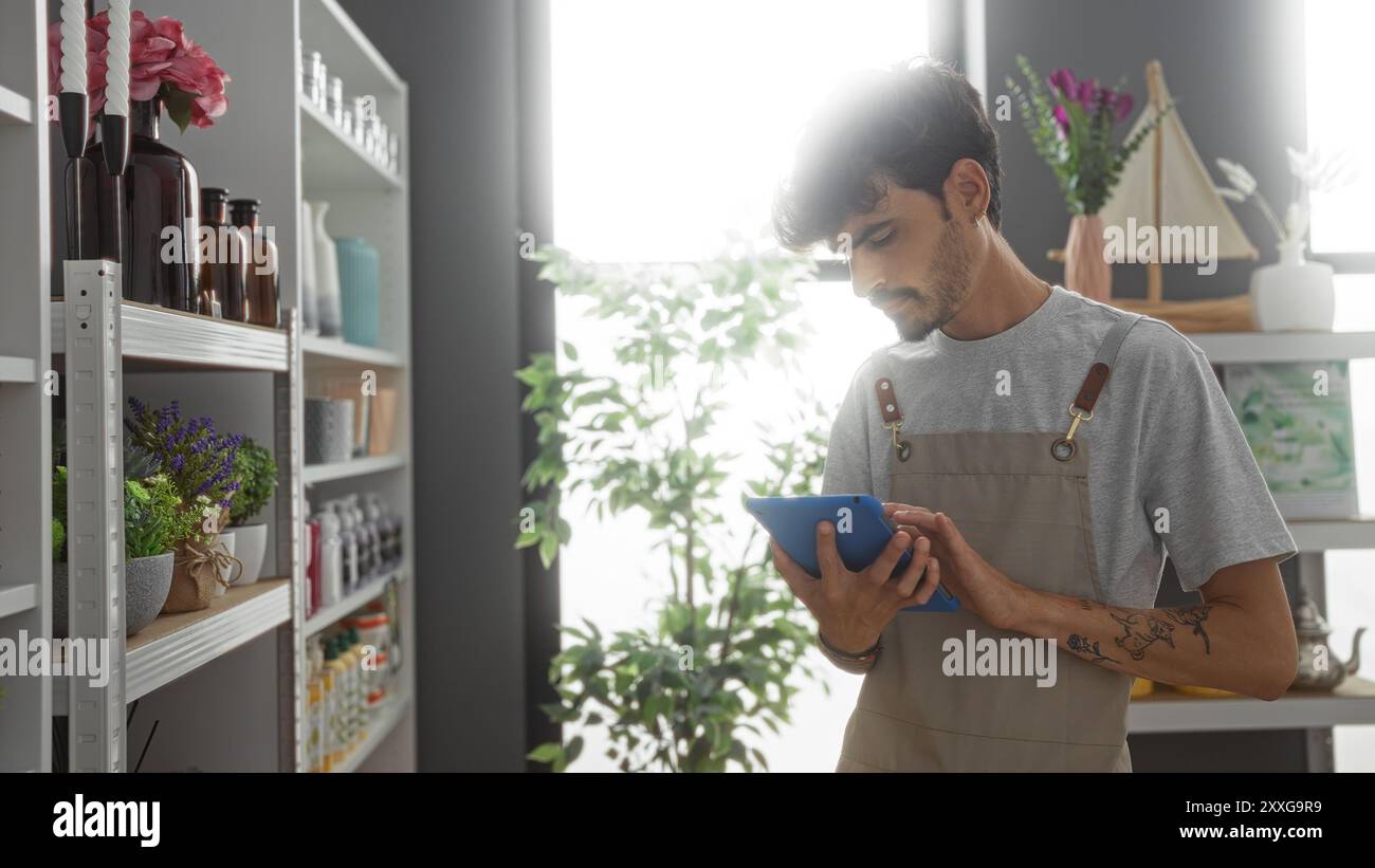 Gutaussehender hispanischer Mann mit Bart im Haus Dekorationsgeschäft mit Tablette für Bestandskontrolle durch Regale mit verschiedenen dekorativen Artikeln unter natürlichem Indoor LIG Stockfoto