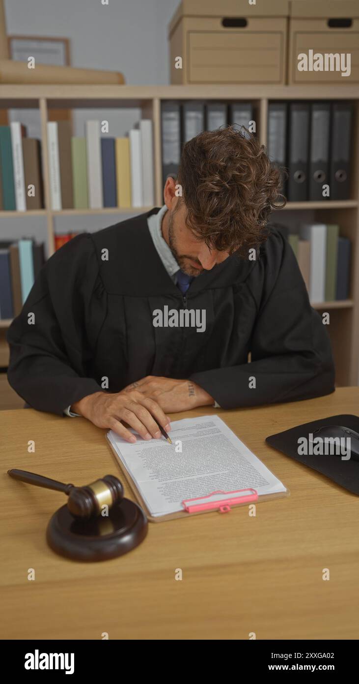 Junger Mann in Richtergewändern, der Dokumente an einem Schreibtisch liest, mit einem Hammer und Bücherregalen im Hintergrund. Stockfoto