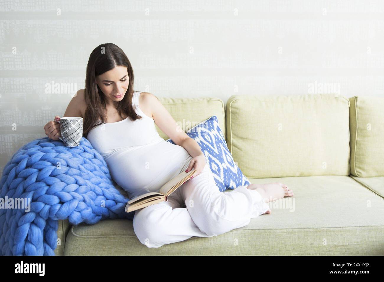 Junge hübsche schwangere Frau drinnen sitzen auf dem Sofa und Lesen Stockfoto