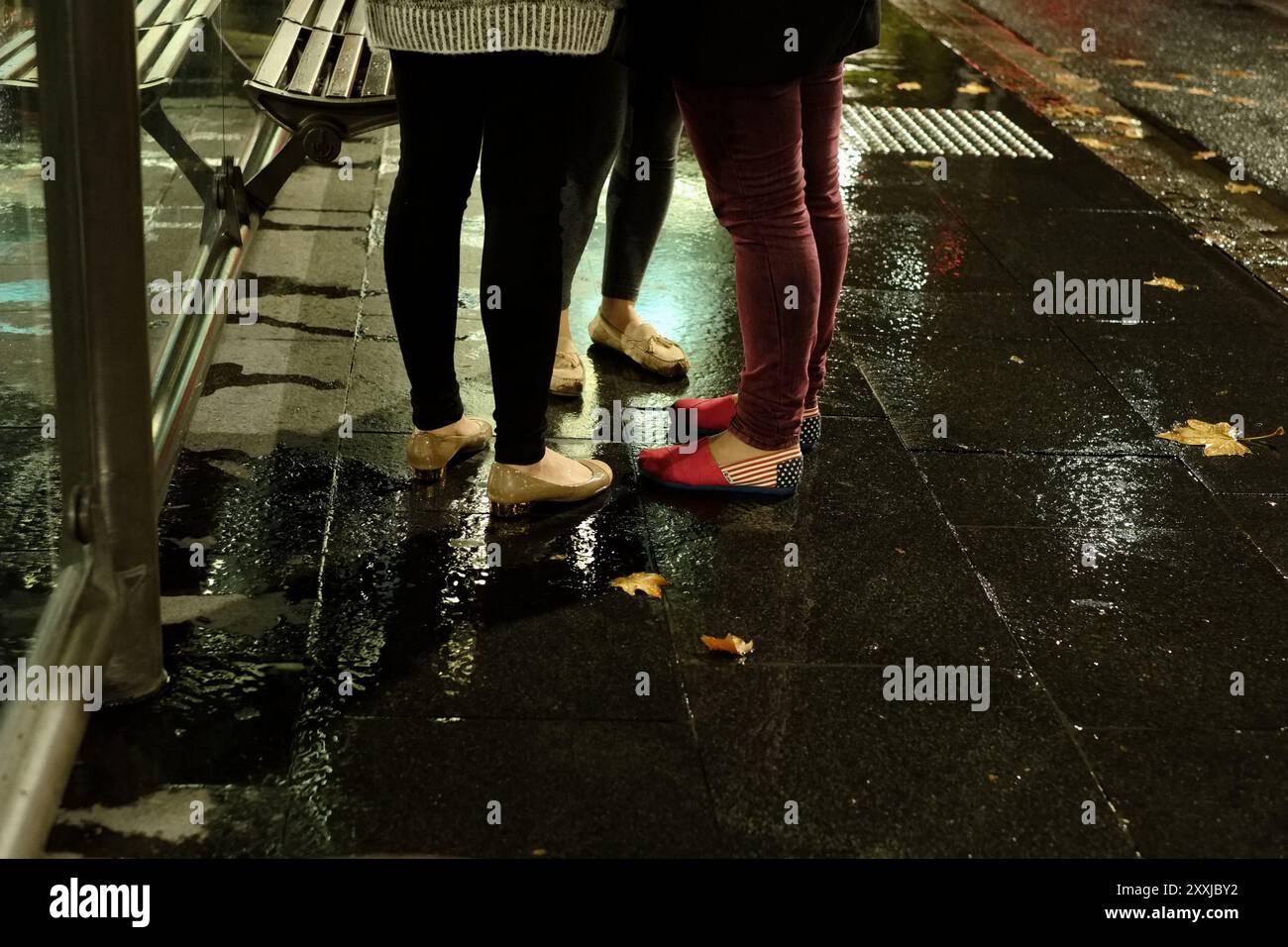 Beine und Schuhe von jungen Frauen, die nachts in einer nassen Bushaltestelle stehen, alle drei tragen Schuhe, ein Paar amerikanische Flagge, Sterne und gestreifte Schuhe Stockfoto