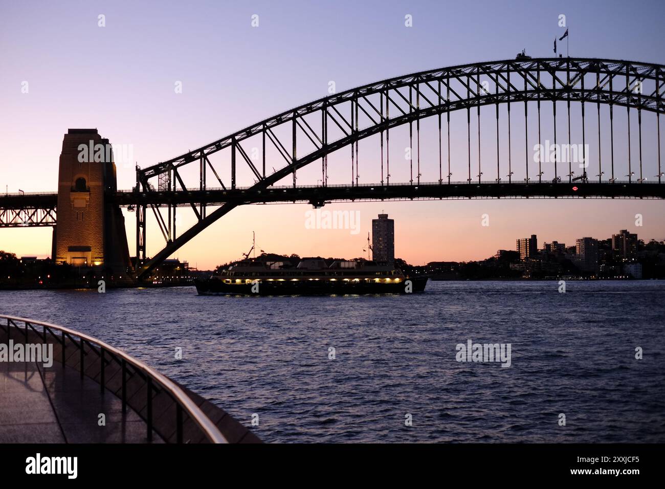 Sydney, Australien, das Art déco-Design der Sydney Harbour Bridge in Silhouette in der Abenddämmerung mit dem Sonnenuntergang hinter dem südlichen Pylon, wenn eine Fähre vorbeifährt. Stockfoto