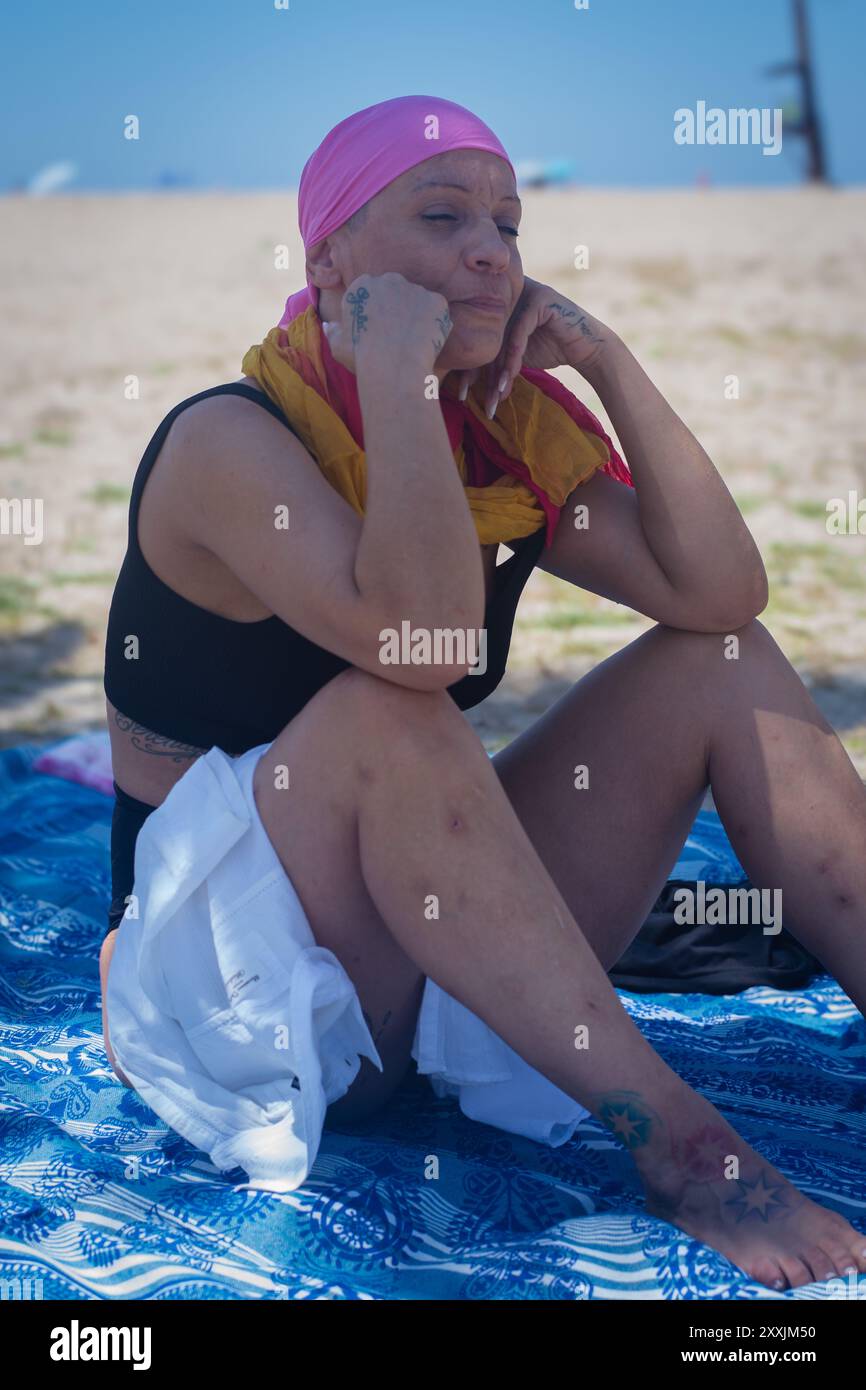 Frau mittleren Alters mit Krebs, die ruhig am Strand sitzt, mit einem kontemplativen Ausdruck unter ihrem rosa Kopftuch Stockfoto