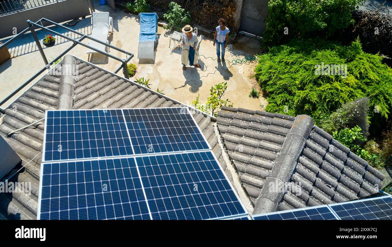 Ein Blick aus der Luft zeigt zwei Frauen in einem sonnigen Hinterhof, eine ist im Garten, während die andere plaudert. Das Dach ist mit Solarpaneelen ausgestattet, Highlight Stockfoto