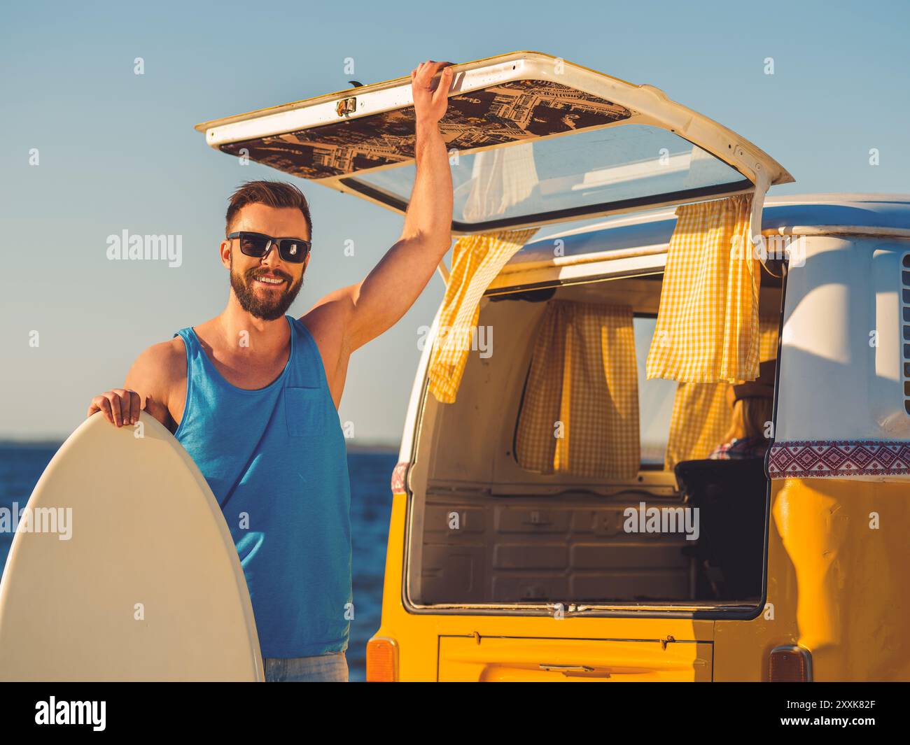 Endlich Sommer! Ein lächelnder junger Mann lehnte sich an das Skimboard und hielt die Hand an der Tür des Kofferraums, während er am Strand stand Stockfoto