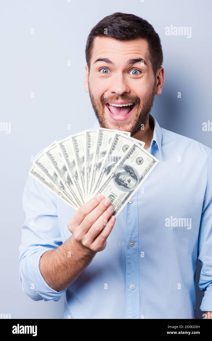 Ich bin reich! Glücklicher junger Mann, der die Papierwährung hält und in die Kamera blickt, während er vor grauem Hintergrund steht Stockfoto