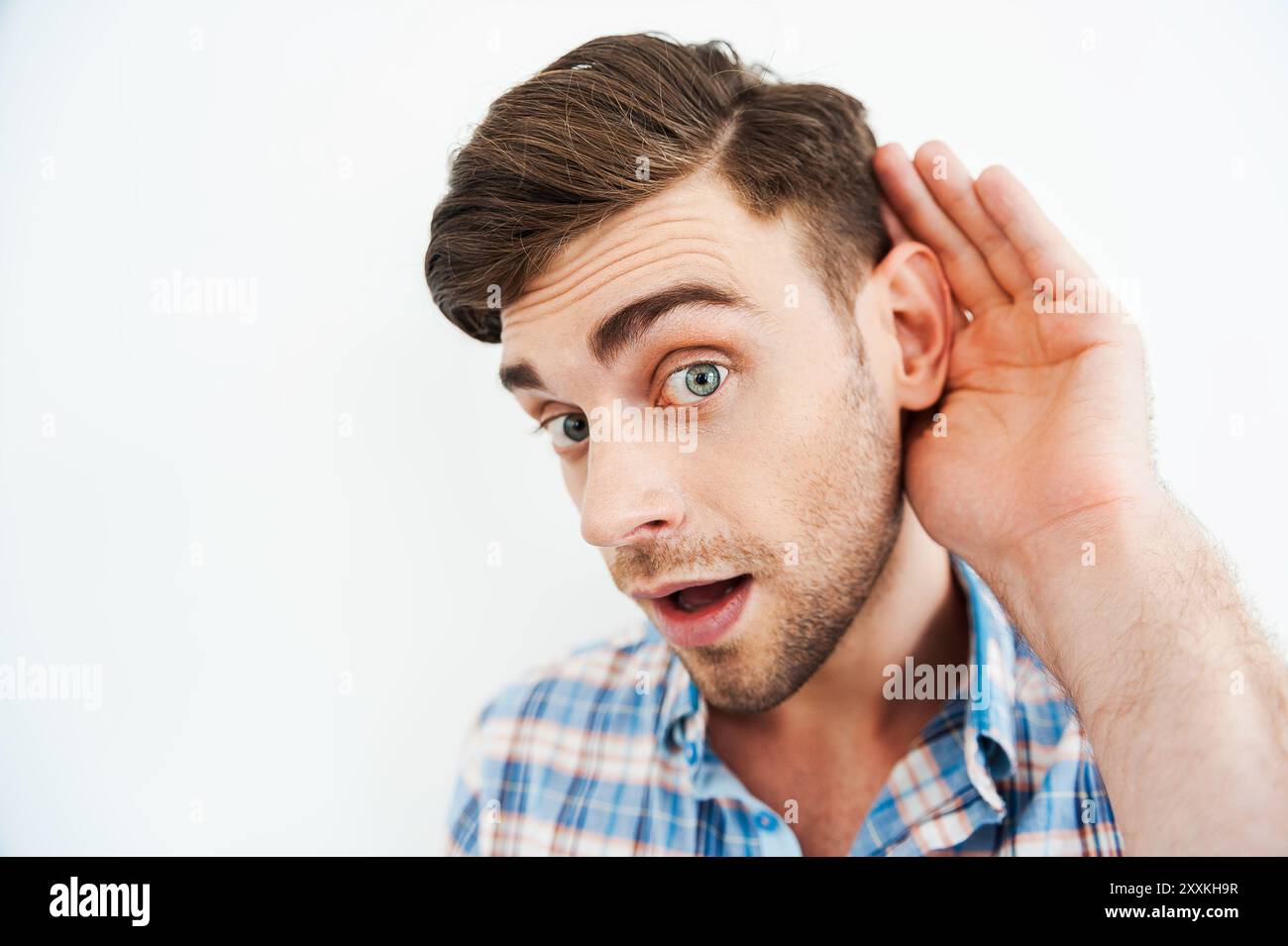 Ich will alles wissen! Neugieriger junger Mann, der Hand hinter Ohr hält und in die Kamera blickt, während er vor weißem Hintergrund steht Stockfoto