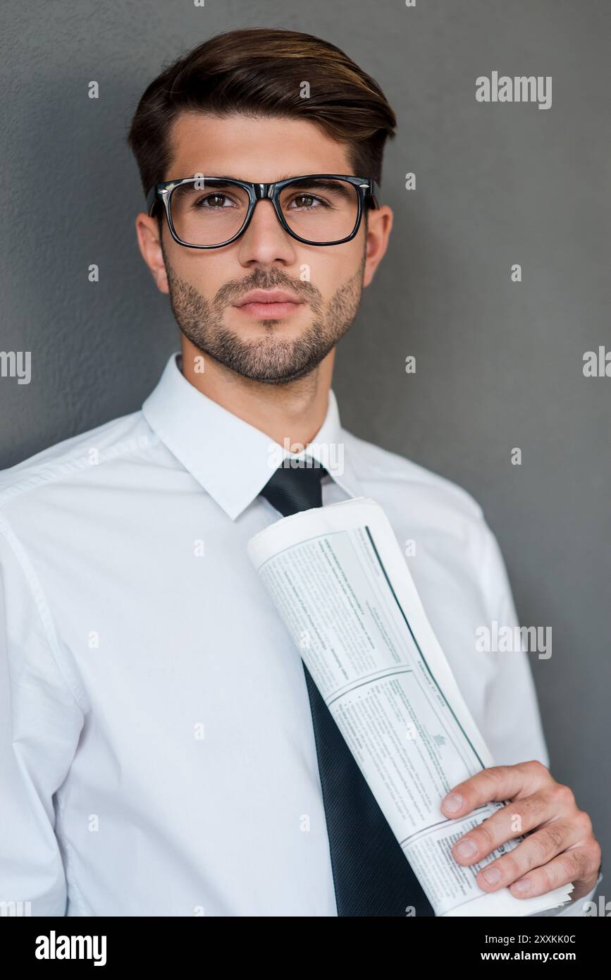 Über neue Lösungen nachdenken. Selbstbewusster junger Mann in Hemd und Krawatte, der Zeitung hält und wegblickt, während er vor grauem Hintergrund steht Stockfoto