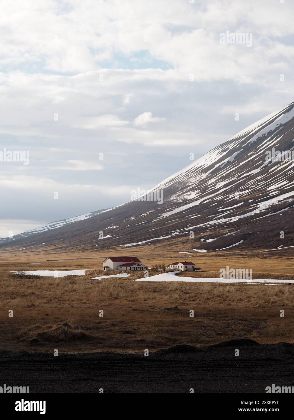 Ein abgelegenes Bauernhaus befindet sich am Fuße eines schneebedeckten Berges unter einem bewölkten Himmel und fängt die ruhige Schönheit des ländlichen und abgelegenen Lebens in ein Stockfoto