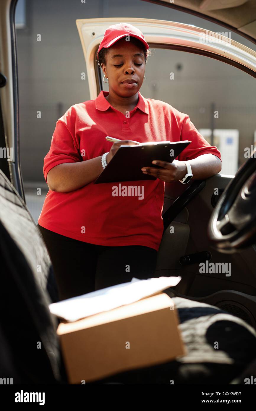 Checkliste des Fahrers, während er in der Fahrzeugtür steht, rote Uniform und Kappe trägt, Clipboard hält, Sonnenlicht durchströmt. Stockfoto