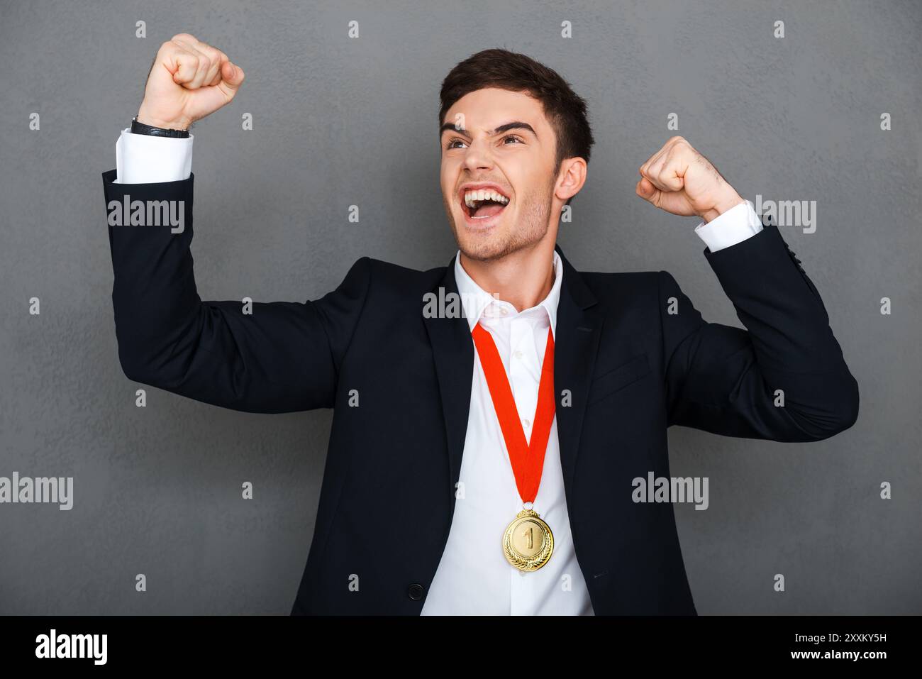 Immer zuerst. Glücklicher junger Mann in formaler Kleidung, der die Arme hoch hält und Positivität ausdrückt, während er vor grauem Hintergrund steht Stockfoto