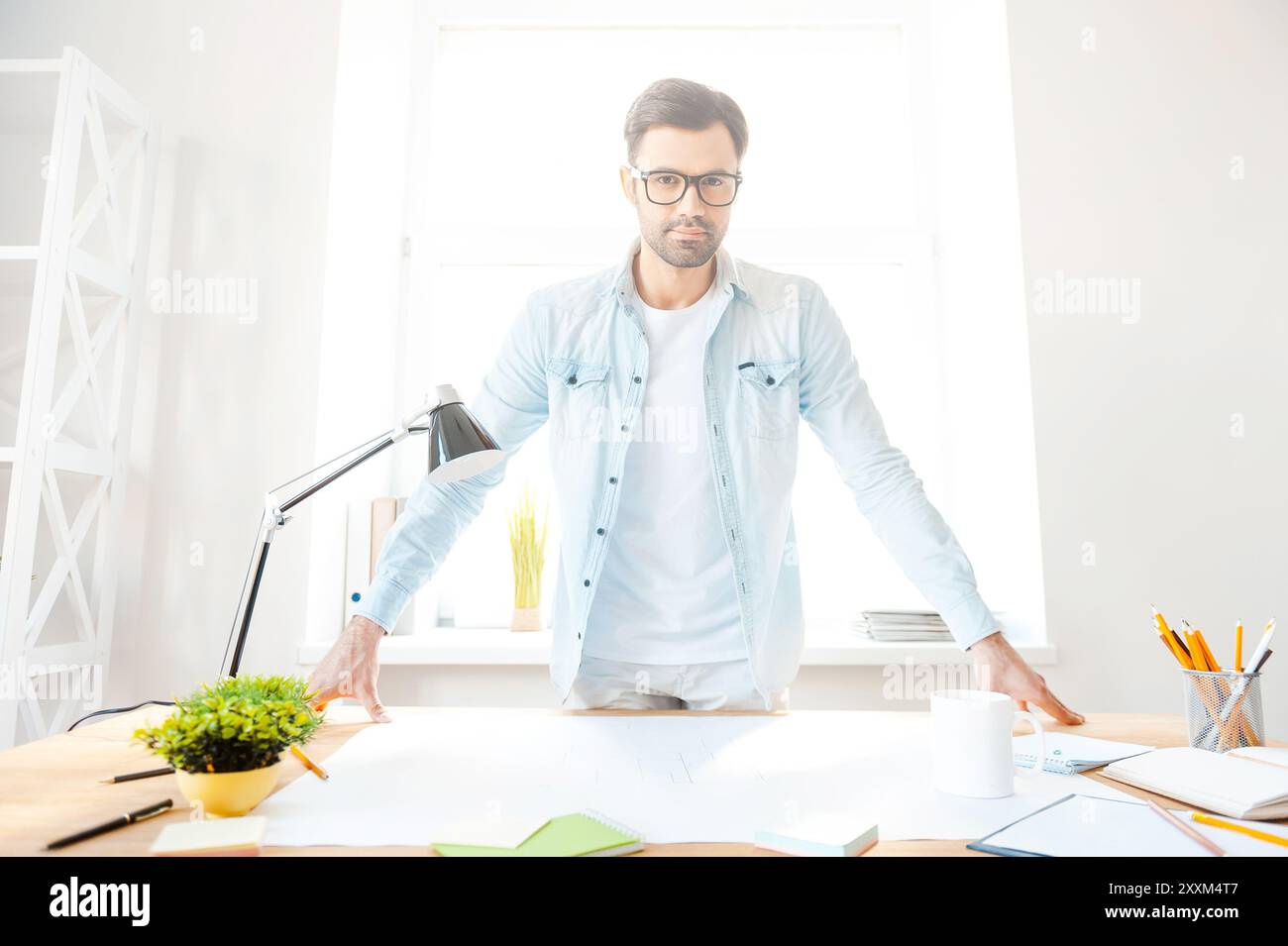 Herr seines Arbeitsplatzes. Hübscher junger Mann in Hemd und Brille, der in die Kamera blickt, während er sich an seinem Arbeitsplatz beugt Stockfoto