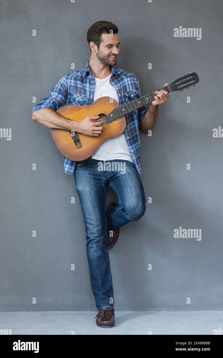 Er zeigt sein musikalisches Talent. Voller fröhlicher junger Mann, der Gitarre spielt und lächelt, während er vor grauem Hintergrund steht Stockfoto