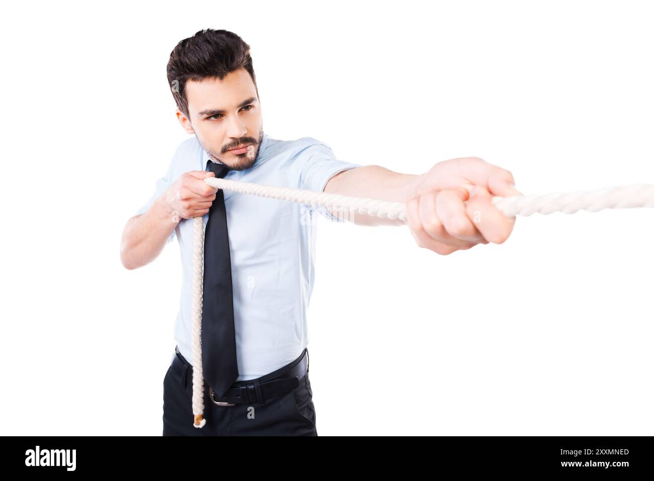 Sieger im Alltag. Selbstbewusster junger Mann in Hemd und Krawatte, der ein Seil zieht, während er vor weißem Hintergrund steht Stockfoto