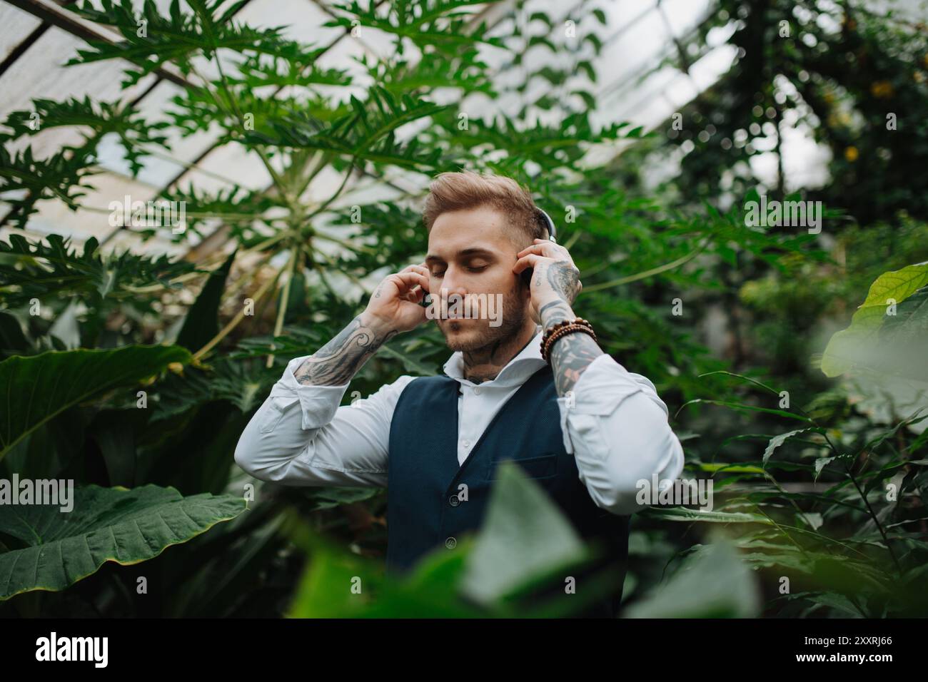 Gutaussehender Mann mit Tätowierung, der Meditationsmusik im botanischen Garten zuhört, inmitten von üppig grünem Laub. Er kümmert sich um seine psychische Gesundheit Stockfoto